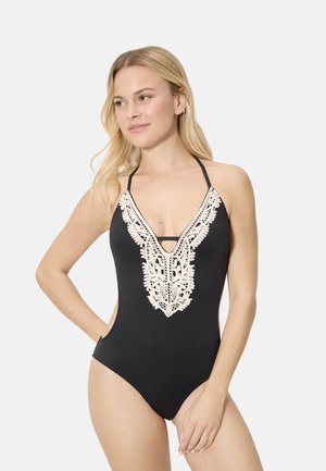 Mujer con bañador negro de cuello halter y escote en V profundo con detalles de encaje blanco, posando con la mano en la cadera frente a un fondo claro.