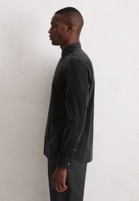 Homme portant une chemise à manches longues noire et un pantalon sombre, se tenant de profil devant un mur blanc uni, regardant vers l'avant.