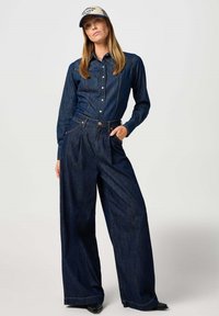 Camisa de mezclilla con cierre de botones, mangas largas y un diseño ajustado, combinada con pantalones de mezclilla oscuros de pierna ancha y cintura alta.