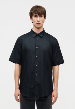 Fiatal férfi rövid barna hajjal, fekete rövid ujjú, finom mintás gombos ingben, egy egyszerű világos háttér előtt áll.