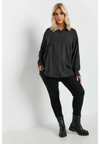 Camisa negra de manga larga con botones al frente, dos bolsillos en el pecho, ajuste ligeramente holgado, combinada con jeans skinny negros y botines negros.