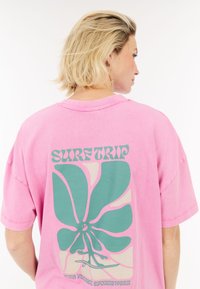 Roze oversized t-shirt met een grote groene bloemgrafiek op de achterkant en de tekst "SURF TRIP". Zachte katoenen stof en een relaxte pasvorm.