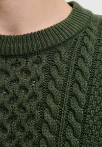 Close-up van een groene gebreide trui met kabelpatronen en een geribbelde ronde halslijn op de hals en schouder van een persoon.