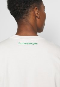 Vit bomull t-shirt med grön text på baksidan som står "det är inte lätt att vara grön." Enkel design med rund hals och korta ärmar.