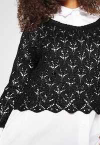 Schwarzer, kurzwolliger Pullover aus gestricktem Stoff, mit einem filigranen Spitzenmuster und einem gewellten Saum. Trägt man über einem weißen Hemd mit Kragen.