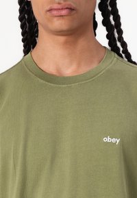 Olive green short-sleeve t-shirt made of cotton, featuring a round neckline and a small white embroidered "obey" logo on the front.
