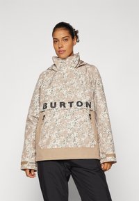 Beżowa kurtka w kwiatowy wzór z wysokim kołnierzem, czarnym logo "BURTON" i kieszeniami na zamek; charakteryzuje się luźnym krojem i teksturowanym materiałem.