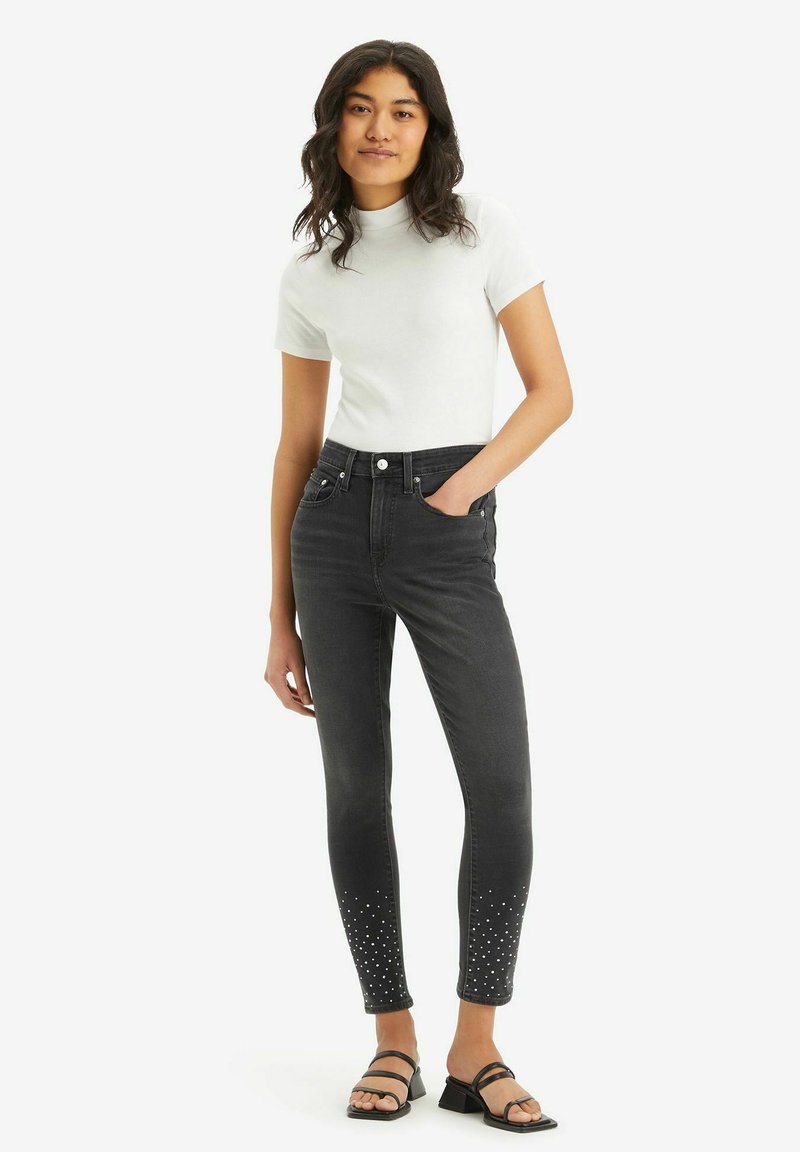 Schwarze Skinny-Jeans mit distressed Details, verziert mit silbernen Nieten am Saum. Getragen mit einem weißen, figurbetonten T-Shirt und schwarzen Sandalen.