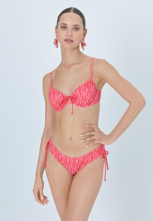 Frau mit zurückgestrichenem Haar, die einen rosa gemusterten Bikini und passende florale Ohrringe trägt, posiert mit einer Hand auf der Hüfte vor einfarbigem Hintergrund.