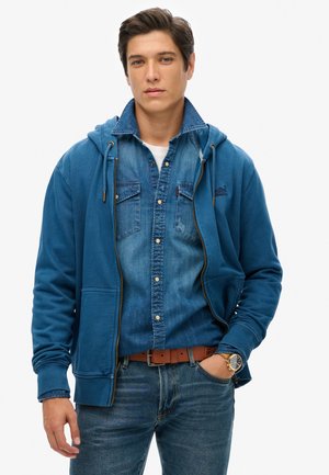 Blå zip-up hættejakke over en lyseblå denimskjorte. Hættejakken har en frontlomme og snører. Mørkeblå jeans og et brunt bælte fuldender looket.