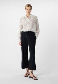 Biała bluzka w kropki z zapięciem na guziki, w połączeniu z czarnymi, szerokimi spodniami i czarnymi sandałami na paski. Gładka tekstura materiału.