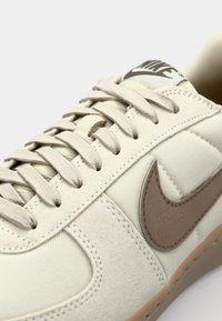 Zapatilla de lona beige con detalles de ante, que presenta un logo Nike en marrón, cordones crema y una suela de goma texturizada. Diseño minimalista.