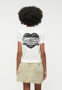 Camiseta blanca de manga corta con un gráfico en forma de corazón negro y el texto "Carhartt Overalls"; combinada con una falda ligera beige con bolsillos.