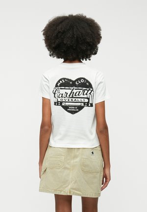 Camiseta blanca de manga corta con un gráfico en forma de corazón negro y el texto "Carhartt Overalls"; combinada con una falda ligera beige con bolsillos.