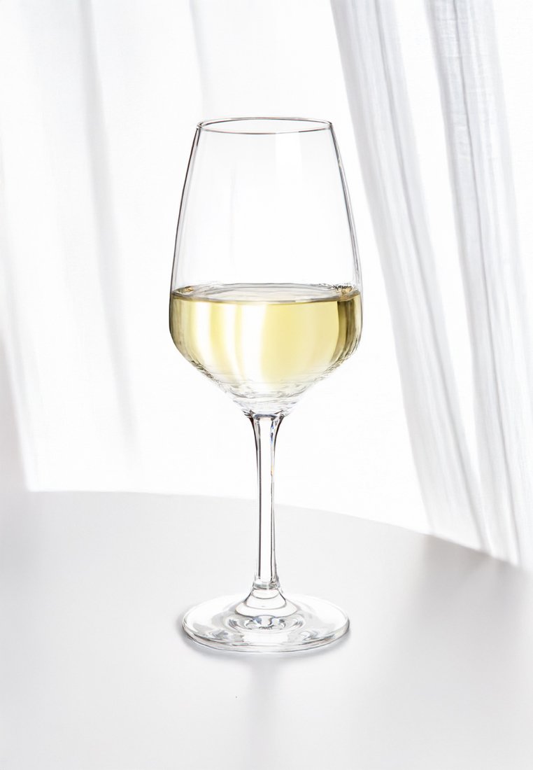 Un verre à vin clair avec une tige élancée, rempli d'un liquide jaune clair, reposant sur une surface blanche circulaire. Une lumière douce provenant d'un rideau léger.