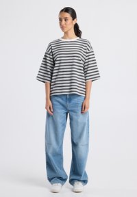 Vrouw staat gekleed in een zwart-wit gestreepte oversized T-shirt, losse lichtblauwe jeans, witte sneakers, tegen een effen lichte achtergrond.