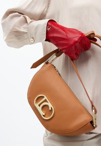 Persona che tiene una borsa in pelle beige con un logo dorato, indossando una blusa chiara e un guanto rosso texturizzato, su uno sfondo semplice.