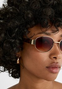Lunettes de soleil avec un cadre en métal doré et des verres teintés brun foncé. Les branches présentent un design écaille de tortue et des accents métalliques distincts.