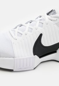 Białe buty sportowe z teksturowanej siateczki i gumowych materiałów. Posiadają czarny logo Nike w kształcie „swoosh” oraz rowkowaną białą podeszwę dla lepszej przyczepności.