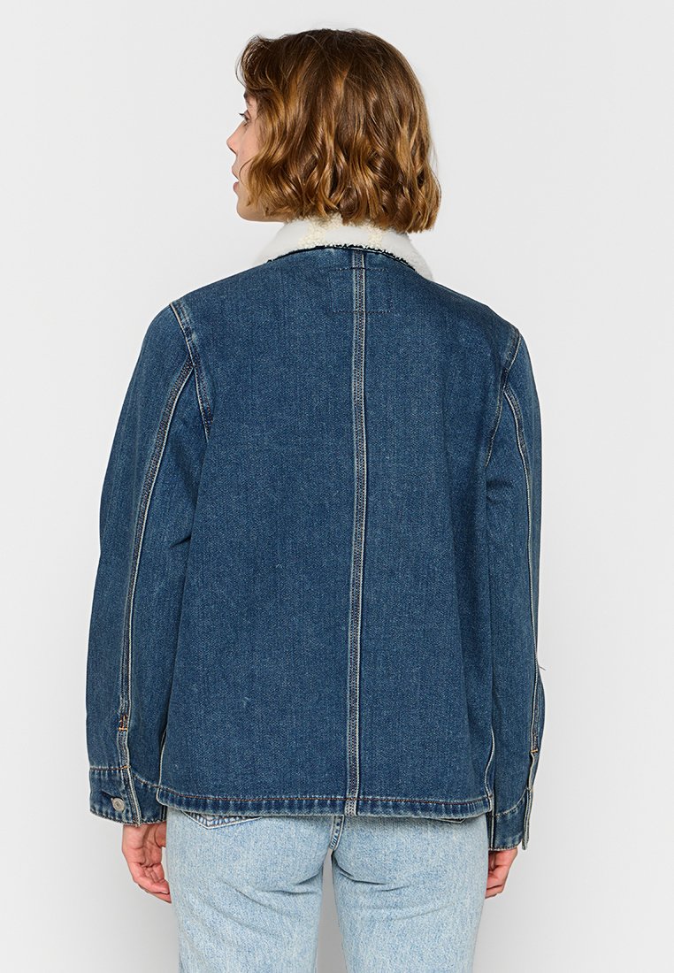 Chaqueta de denim en azul oscuro con un cuello de borreguito blanco y costuras de contraste. Presenta un corte holgado y dos bolsillos laterales.