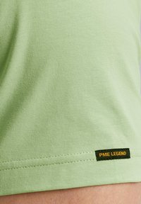Camiseta de algodón verde claro con una textura suave; presenta una etiqueta PME LEGEND en negro y amarillo en el dobladillo. Diseño de manga corta.