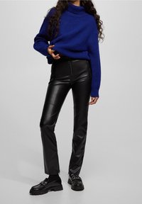 Pull en tricot bleu à col roulé et pantalon noir en simili cuir avec poches zippées. Chaussures avec semelle épaisse et accents métalliques.