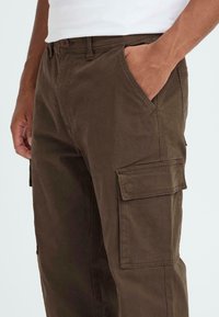 Braune Cargo-Hosen aus Baumwolle mit weicher Textur, ausgestattet mit Seitentaschen und einem Knopfverschluss. Die Passform ist lässig mit sichtbaren Nähdetails.