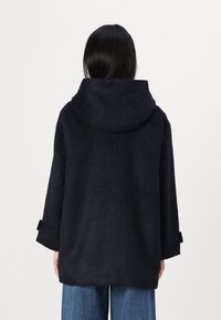 Persona con cabello largo y negro, vistiendo un abrigo holgado con capucha de color azul marino oscuro y jeans anchos azules, de pie con la espalda hacia el espectador.
