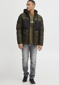 Vadderad olivgrön jacka med en svart bröstpanel, dragkedjefickor och huva, i kombination med en grafisk olivgrön t-shirt och grå jeans.