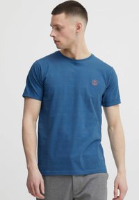 Tricou scurt cu mânecă, de culoare albastră, confecționat dintr-un material moale cu o textură heathered. Prezintă un mic logo portocaliu pe partea stângă a pieptului. Guler rotund.
