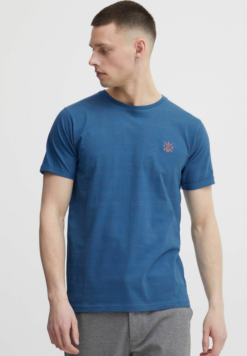 Tricou scurt cu mânecă, de culoare albastră, confecționat dintr-un material moale cu o textură heathered. Prezintă un mic logo portocaliu pe partea stângă a pieptului. Guler rotund.