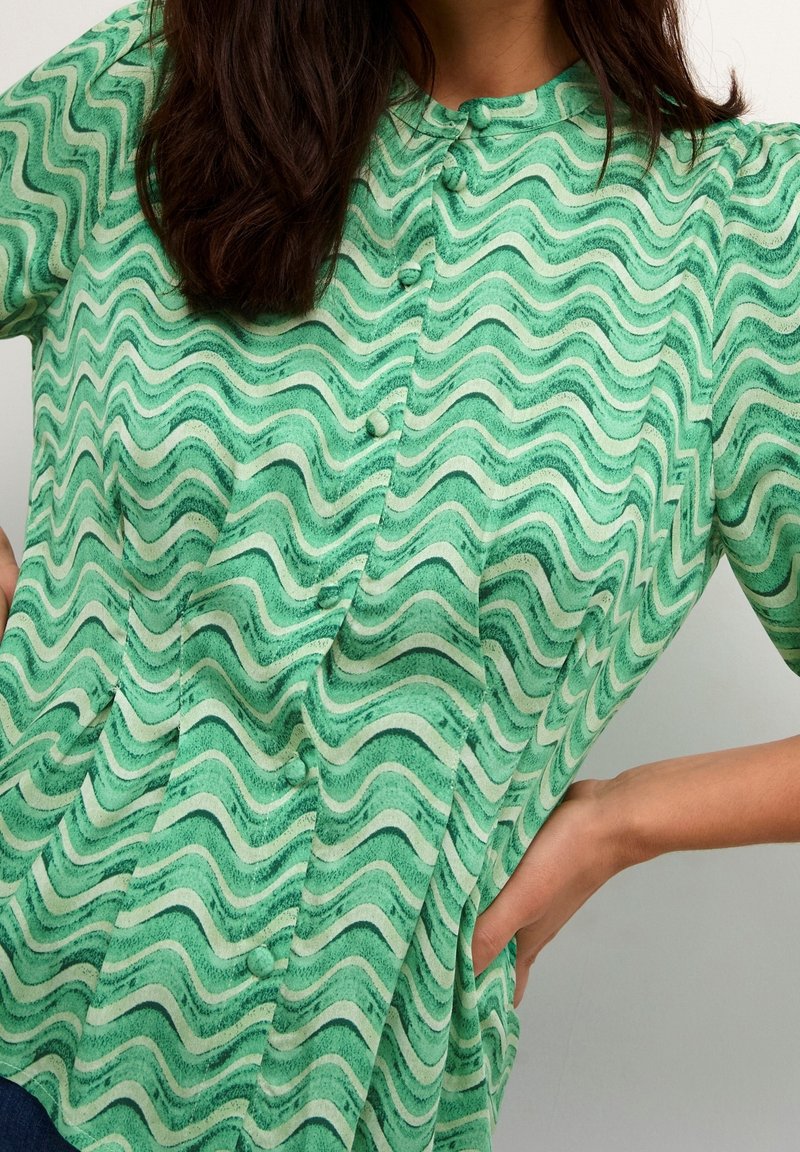 Donna che indossa una camicia verde a motivi ondulati con una mano appoggiata sull'anca contro uno sfondo semplice.