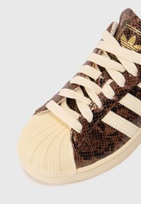 Sapatilha com um padrão de pele de cobra castanha, biqueira de borracha bege e atacadores de cor creme. Apresenta três riscas brancas de lado e detalhes texturizados.