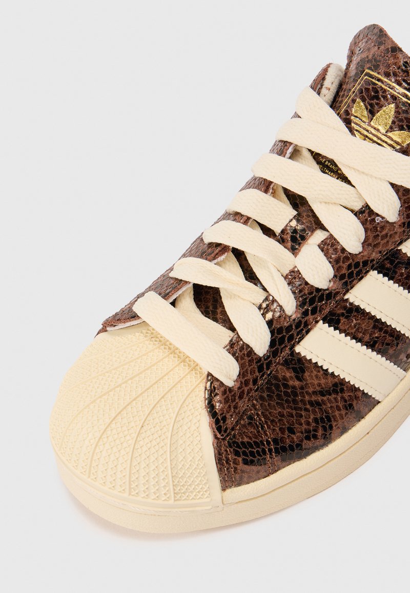 Basket avec un motif de peau de serpent brun, un capuchon en caoutchouc beige et des lacets crème. Comprend trois stripes blanches sur le côté et des détails texturés.