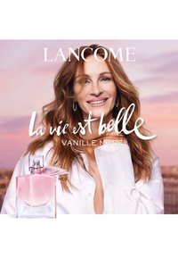 Rosa glassparfymeflaske med rund form, sølvdetaljer og et rosa bånd. Teksten sier "La vie est belle Vanille Nude."