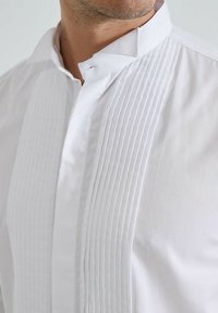 Chemise blanche avec un col mandarin et des plis verticaux à l'avant. Fabriquée en tissu lisse avec une finition nette et un matériel minimal.