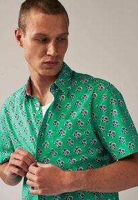 Grünes Kurzarmhemd mit Blumenmustern in Pink und Weiß. Verfügt über einen Button-Down-Kragen und subtile Streifenakzente am inneren Kragen.