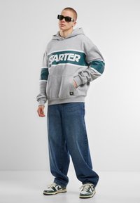 Hoodie gris avec logo "STARTER" en blanc et turquoise. Associé à un jean bleu ample et des baskets vertes et blanches au design classique.