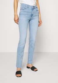 Vaqueros skinny de un azul claro, fabricados en mezclilla, con una cintura de altura media, diseño de cinco bolsillos y dobladillo deshilachado. Combinados con sandalias negras de tiras tejidas.