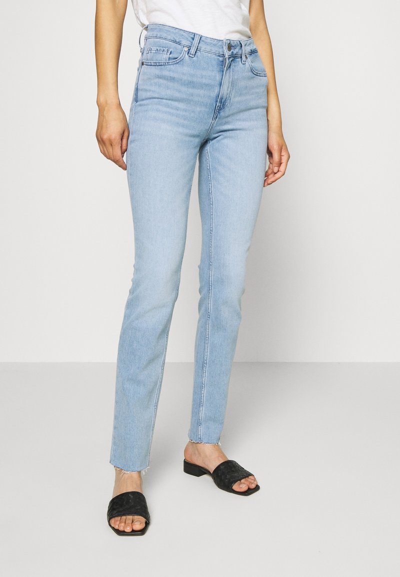 Vaqueros skinny de un azul claro, fabricados en mezclilla, con una cintura de altura media, diseño de cinco bolsillos y dobladillo deshilachado. Combinados con sandalias negras de tiras tejidas.