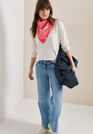 Femme portant une écharpe rouge à motifs, un haut à manches longues crème, un jean bleu clair, des baskets blanches et tenant un grand sac cabas bleu marine.