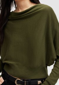 Olivgrüner Pullover mit einem drapierten Kragen und langen Ärmeln, der ein strukturiertes Strickmuster aufweist. Kombiniert mit einem schwarzen Gürtel, der eine goldene Schnalle präsentiert.