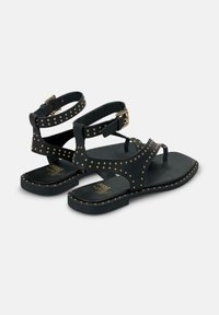 Sandalias de cuero negro con tachuelas doradas, dos correas en el tobillo y suela plana. Presenta un lazo para el dedo del pie y cierre de hebilla.