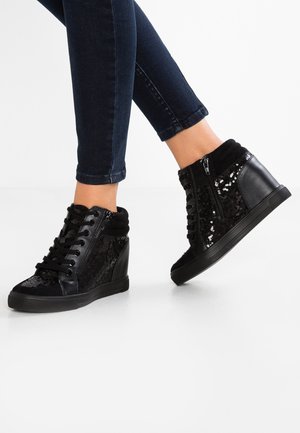 Person iført sorte kilehælssneakers med pailletter og lynlåse, kombineret med mørke skinny jeans, stående på et hvidt gulv.