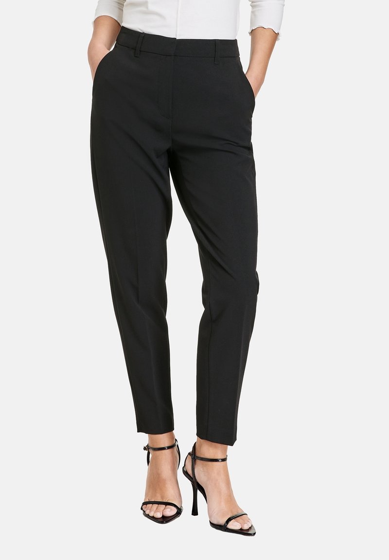 Femme portant un pantalon noir ajusté à longueur de cheville avec des poches, associé à des sandales à talons hauts noires à lanières et un haut blanc.