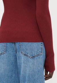Burgundy bordázott kötött pulóver ujja magas derekú kék farmernadrággal, amely folt zsebekkel és elhalványult textúrával rendelkezik.