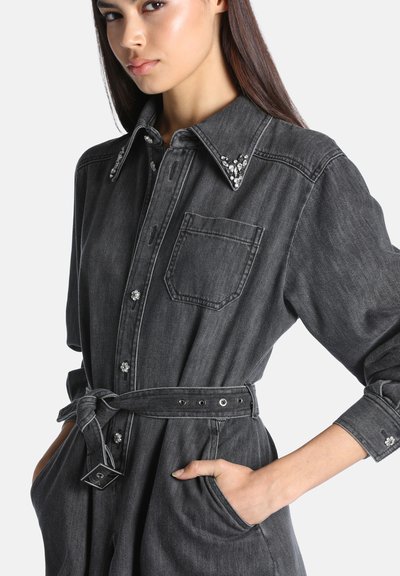 Femme portant une robe en denim gris foncé ceinturée avec des boutons et un col ornés, les mains dans les poches, légèrement tournée vers la gauche sur un fond uni.