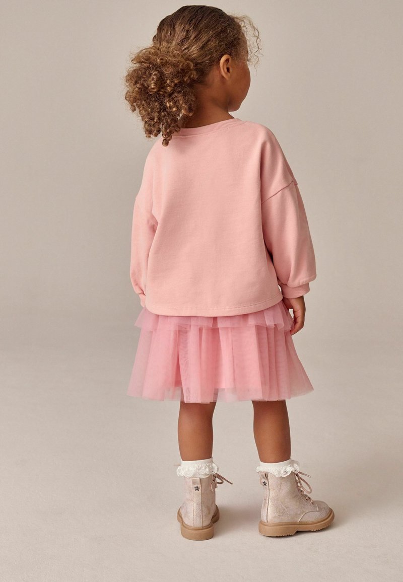 Sweatshirt rosa, gonna in tulle sovrapposta di un rosa chiaro e stivaletti alla caviglia lucidi di un rosa chiaro con dettagli in pizzo bianco. Capelli ricci visibili.