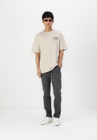 Camiseta oversized de color beige claro con estampado gráfico, combinada con pantalones tapered de color gris oscuro y zapatillas blancas, llevada por un modelo con gafas de sol.