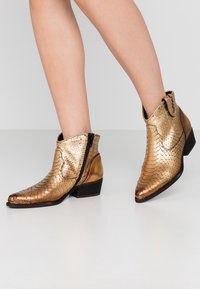 Gold ankle boots with a textured, scaly design, featuring a side zipper and low block heel. Pointed toe and shiny finish.